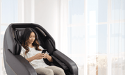 A woman sits in a Daiwa Hubble Massage Chair, using the phone, showcasing its compact, wall-hugging design and relaxing features.